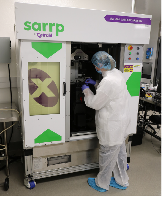 A researcher in protective clothing operates a SARRP machine. The individual is wearing a white lab coat, blue hair cover, face mask, purple gloves, and blue shoe covers. The machine features a door with green triangular designs and an 'X' symbol, along with buttons and warning signs on its side.